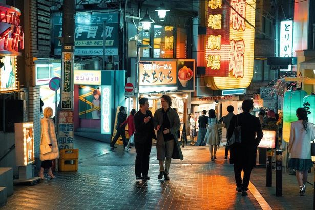 ネオンがきらめく東京の繁華街を練り歩くアンセル・エルゴート演じるジェイクと伊藤英明扮する刑事の宮本